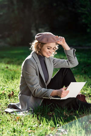 Work outdoors benefits. Education technology and internet concept. Smiling girl in a beret work with laptop in park sitting on grass. Natural environment office.の写真素材