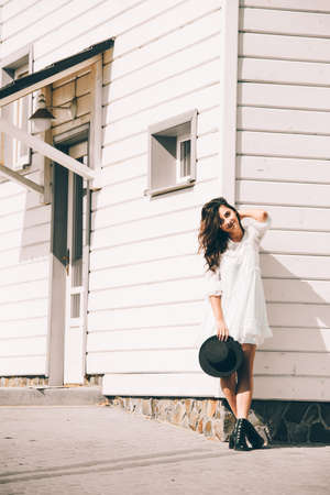 Sunny lifestyle fashion portrait of young stylish hipster woman walking on the street, wearing trendy white dress, black hat and boots. White wooden house on a backgrond.の写真素材