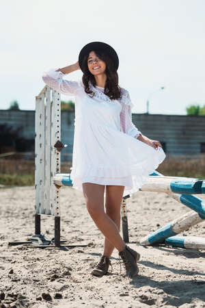 Beautiful sexy girl posing on the horizontal crosspiece on a horse epodrom. Female wearing white dress, black hat and black boots. Summertimeの写真素材