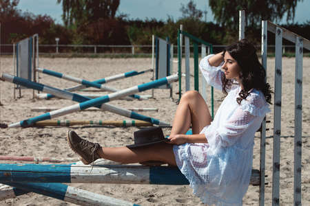 Beautiful sexy girl posing on the horizontal crosspiece on a horse epodrom. Female wearing white dress, black hat and black boots. Summertimeの写真素材