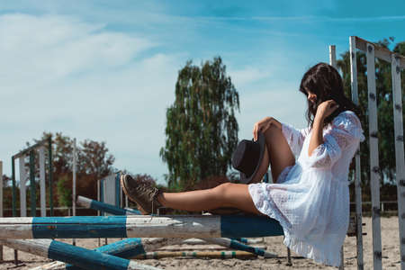 Beautiful sexy girl posing on the horizontal crosspiece on a horse epodrom. Female wearing white dress, black hat and black boots. Summertimeの写真素材