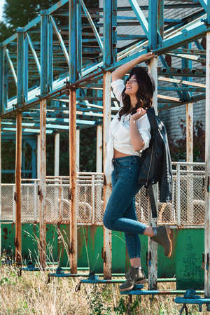 Beautiful sexy girl posing on the rustic metal construction. Female wearing white shirt, black hat and blue denim jeans. black jacket on the shoulderの写真素材