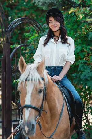 Portrait of young brunette woman in a white shirt and helmet on a horse. learns to ride a horse. riding lessons in a parkの写真素材