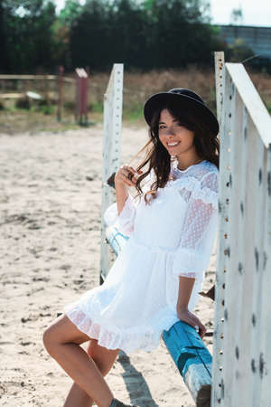 Beautiful sexy girl posing on the horizontal crosspiece on a horse epodrom. Female wearing white dress, black hat and black boots. Summertimeの写真素材