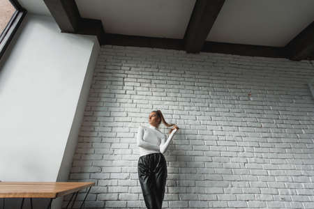 Fashionable brunette girl in a white sweater and black leather pants posing on an white bricks...