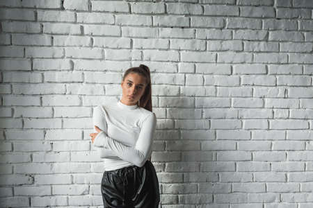 Fashionable brunette girl in a white sweater and black leather pants posing on an white bricks background.の写真素材