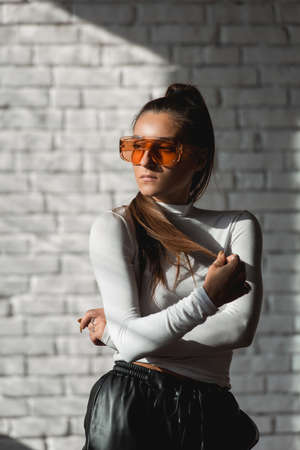 Fashionable brunette girl in a white sweater, orange glasses and black leather pants posing on an white bricks background.の写真素材