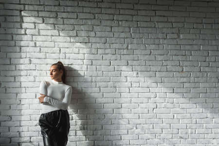 Fashionable brunette girl in a white sweater and black leather pants posing on an white bricks background.の写真素材