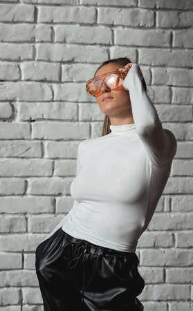 Fashionable brunette girl in a white sweater, orange glasses and black leather pants posing on an white bricks background.の写真素材