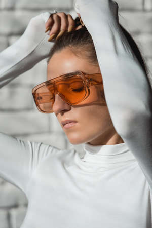 Fashionable brunette girl in a white sweater, orange glasses and black leather pants posing on an white bricks background.の写真素材