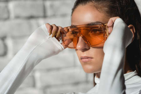 Fashionable brunette girl in a white sweater, orange glasses and black leather pants posing at the sunshine on an white bricks background.の写真素材