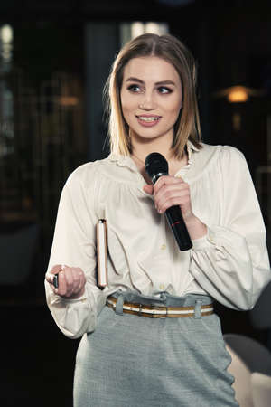 Business and speech topic: woman in a white blouse holding a black microphone on a dark backgroundの写真素材