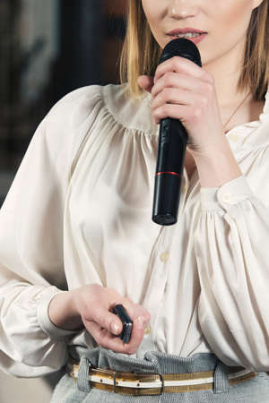 Business and speech topic: woman in a white blouse and gray skirt holding a black microphone in one hand and a clicker in another, indoorの写真素材