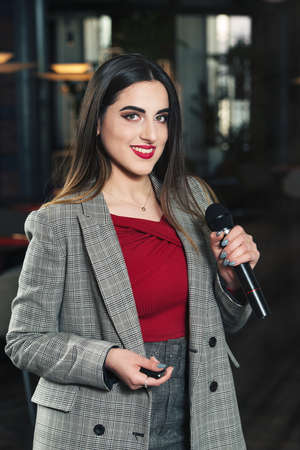 Business and speech topic: woman in a red shirt and gray jacket holding a black microphone in one hand and a clicker in another, indoorの写真素材