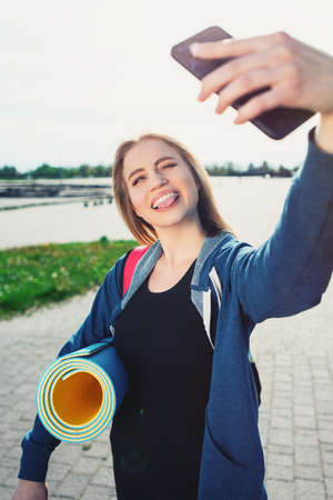 young beautiful smiling sport girl in sports clothes outdoors after a workout and making selfie on a smart phone. Healthy lifestyle concept.の写真素材