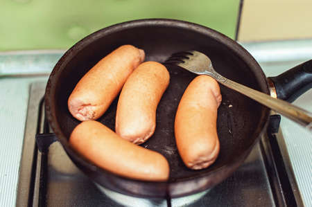 Close up view of sausages being fried with cooking oil in a pan. Pan is on a gas stove with background of tiled kitchen wall. Selective focusの写真素材