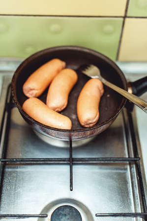 Close up view of sausages being fried with cooking oil in a pan. Pan is on a gas stove with background of tiled kitchen wall. Selective focusの写真素材