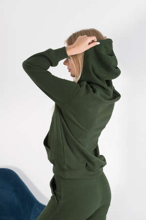 Stylish beautiful young blond woman in a green tracksuit poses near a white wall in the room. Attractive girl model posing near blue chair.の写真素材