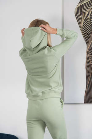 Stylish beautiful young blond woman in a green tracksuit poses near a white wall in the room.の写真素材