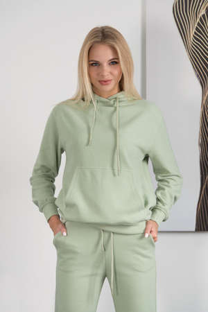 Stylish beautiful young blond woman in a green tracksuit poses near a white wall in the room.の写真素材