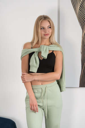 Stylish beautiful young blond woman in a green tracksuit poses near a white wall in the room.の写真素材