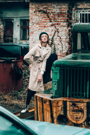 Beautiful young stylish blonde woman wearing long beige coat, white boots, black hat, umbrella and backpack posing through the city streets. Trendy casual outfit. Selective focus, grainの写真素材