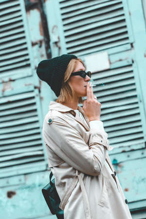 Beautiful young stylish blonde woman wearing long beige coat, white boots, black hat, umbrella and backpack posing through the city streets. Trendy casual outfit. Selective focus, grainの写真素材