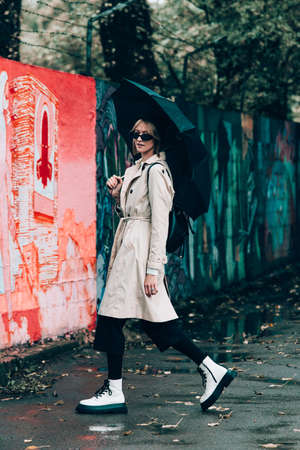 Beautiful young stylish blonde woman wearing long beige coat, white boots, black hat, umbrella and backpack posing through the city streets. Trendy casual outfit. Selective focus, grainの写真素材