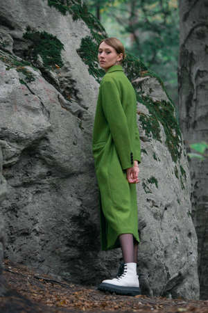 Fashionable young woman in green coat big white boots and black tights posing in a forest sitting on a stone. Art portraitの写真素材