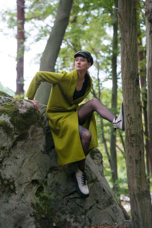 Fashionable young woman in green coat big white boots and black tights posing in a forest sitting on a stone. Art portraitの写真素材
