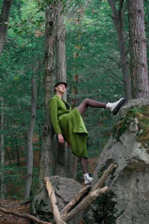 Fashionable young woman in green coat big white boots and black tights posing in a forest sitting on a stone. Art portraitの写真素材