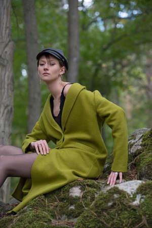 Fashionable young woman in green coat, cap and black tights posing in a forest on a stone. Art portraitの写真素材