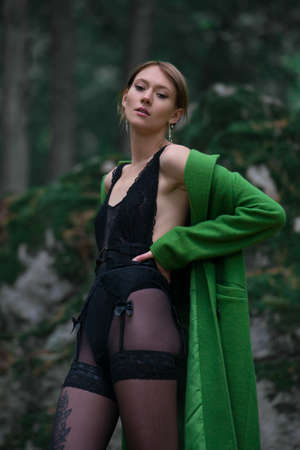 Fashionable young woman in green coat and black tights posing in a forest sitting on a stone. Art portraitの写真素材