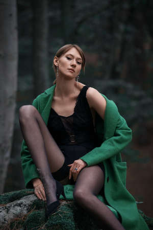 Fashionable young woman in green coat black tights, leather heeled shoes posing in a forest sitting on a stone. Art portraitの写真素材
