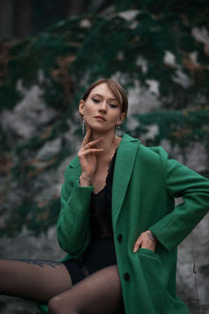 Fashionable young woman in green coat and black tights posing in a forest sitting on a stone. Art portraitの写真素材
