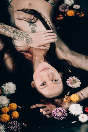 Young pretty brunette woman with tattoos in bath with water and flowers. Naked model during an unusual photo shoot in a black bath.の写真素材