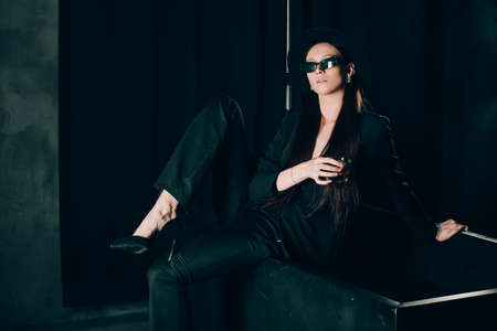 girl in a black trouser suit and hat posing near a black bath. A glass with alcohol in hand. Stylish sunglassesの写真素材