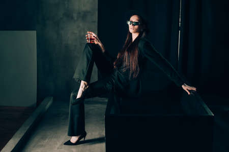girl in a black trouser suit and hat posing near a black bath. A glass with alcohol in hand. Stylish sunglassesの写真素材