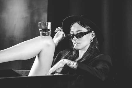 girl in a black jacket and hat smoking cigarette posing near a black bath. A glass with alcohol in hand. Stylish sunglasses. Long naked sexy legsの写真素材