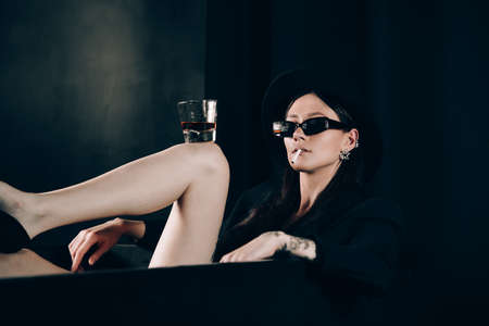girl in a black jacket and hat smoking cigarette posing near a black bath. A glass with alcohol in hand. Stylish sunglasses. Long naked sexy legsの写真素材