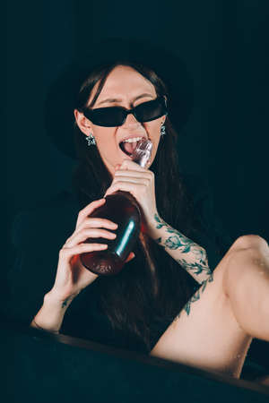 girl in a black jacket and hat posing in a black bath. A bottle of rose champagne wine in hand. Stylish sunglasses. Long naked sexy legsの写真素材
