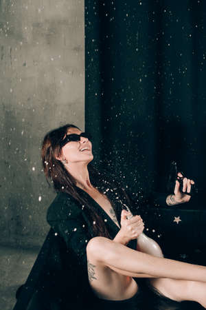 girl in a black jacket posing in a black bath. A bottle of rose champagne wine in hand. Stylish sunglasses. Long naked sexy legs. splashes of champagneの写真素材