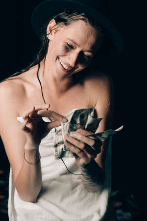 Cheerful attractive young lady in a towel and a hat holding cash and happy smiling over black backgroundの写真素材