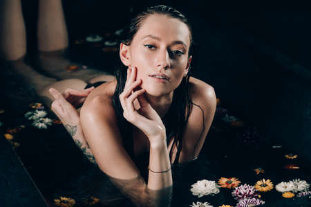 Young pretty brunette woman with tattoos in bath with water and flowers. Naked model during an unusual photo shoot in a black bath.の写真素材