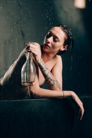 Young pretty brunette woman with tattoos in a bath. Naked model during an unusual photo shoot in a black bath drinking wineの写真素材