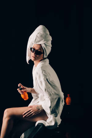 Skin Care, Woman in Sunglasses Sitting on the Edge of Black Bath Applying Cream in Towel after Spa, Treatment Cosmetic Moisturizing Lotion. Black backgroundの写真素材