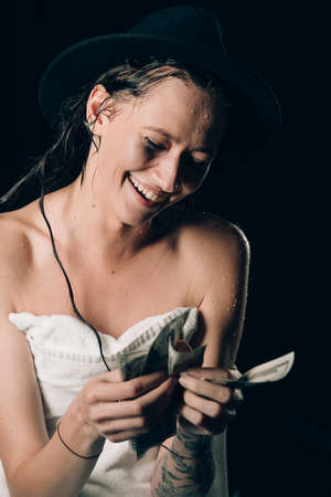 Cheerful attractive young lady in a towel and a hat holding cash and happy smiling over black backgroundの写真素材