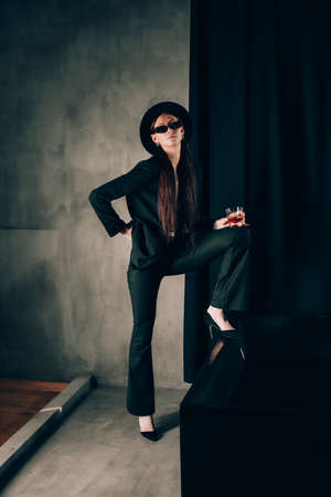 girl in a black trouser suit and hat posing near a black bath. A glass with alcohol in hand. Stylish sunglassesの写真素材