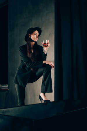girl in a black trouser suit and hat posing near a black bath. A glass with alcohol in hand. Stylish sunglassesの写真素材