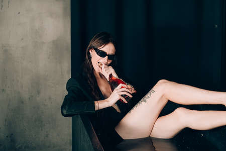 girl in a black jacket in a black bath. Opening a bottle of rose champagne wine. Stylish sunglasses. Long naked sexy legsの写真素材